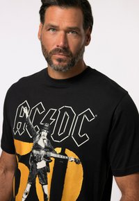 Sort bomulds T-shirt med et grafisk print af en rockmusiker med en elektrisk guitar, fremhævet af fed hvid og gul skrift.