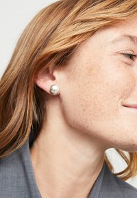 Pendientes de perla plateada en forma de botón, forma redonda, superficie lisa, tamaño mediano, llevados en una oreja de piel clara, con pecas visibles y cabello castaño.