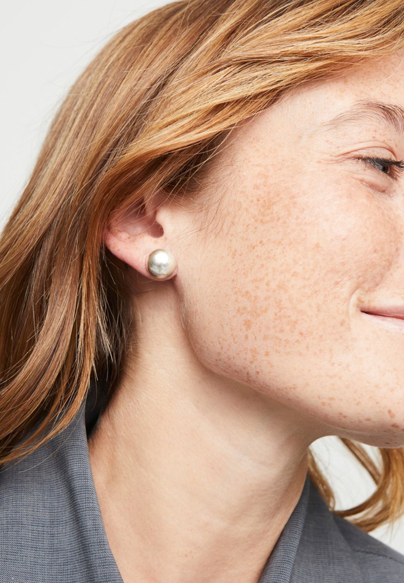 Pendientes de perla plateada en forma de botón, forma redonda, superficie lisa, tamaño mediano, llevados en una oreja de piel clara, con pecas visibles y cabello castaño.