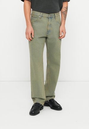 Mężczyzna noszący wyblakłe zielone jeansy o prostych nogawkach, czarne skórzane loafersy oraz czarną koszulę z krótkim rękawem, z widocznym tatuażem na lewym ramieniu.