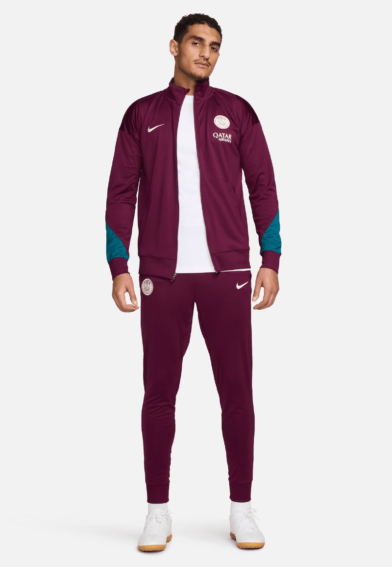 Maroon urheilutakki ja housut, joissa on teal-väriä aksentteina, korkea kaulus, rintalogo ja valkoiset lenkkarit. Materiaali on sileä ja kevyt.