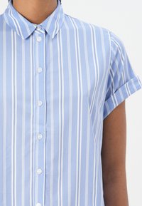 Svetlomodrá košeľa s krátkymi rukávmi a vertikálnymi bielymi pruhmi, s button-up zapínaním a klasickým golierom. Mäkká bavlnená tkanina s jemnou textúrou.