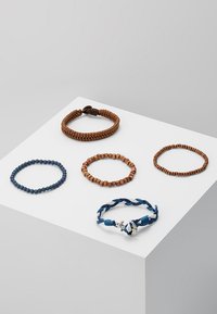Cinq bracelets : deux en cordon marron, un avec une perle bleue, un avec une perle marron, et un en tressé bleu et blanc avec un accent en argent.