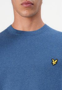 Pull bleu à col rond en tissu tricoté doux. Présente un petit emblème d'oiseau brodé jaune sur la poitrine gauche.