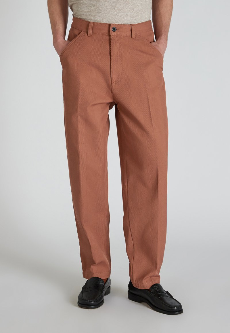 Uomo in pantaloni color ruggine con le mani in tasca e mocassini neri, in piedi contro uno sfondo chiaro e semplice.