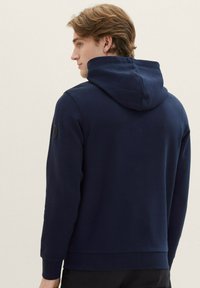 Felpa blu scuro con superficie testurizzata, cappuccio regolabile, polsini a coste e dettaglio con logo rotondo sulla spalla sinistra.