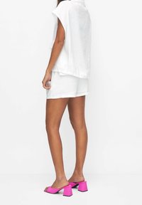 Person som bär en vit ärmlös topp och shorts, ihop med klara rosa klackskor, som står mot en enkel vit bakgrund.