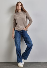 Vaalea ruskea pitkähihaisen paita, jossa on korkea kaula-aukko, yhdistettynä sinisiin leveisiin farkkuihin ja valkoisiin tennareihin, neutraalia taustaa vasten.