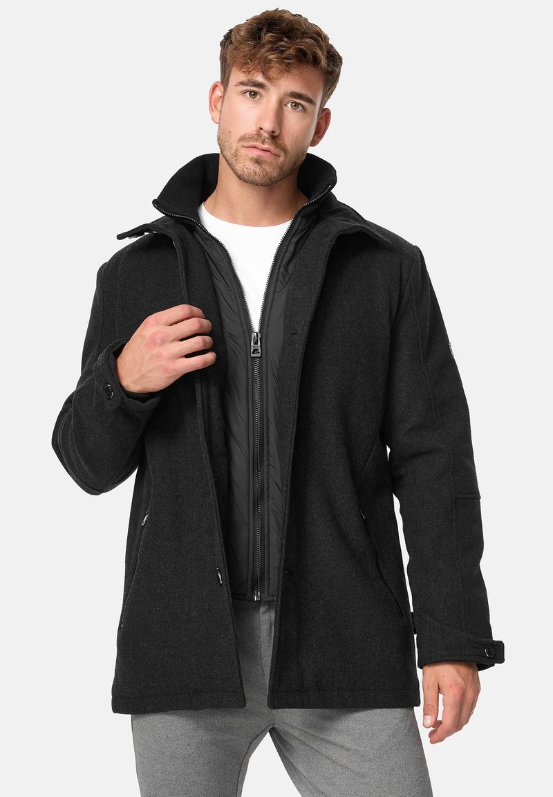 Chaqueta de mezcla de lana negra con una capa interior negra removible, cierre de cremallera, bolsillos laterales y un collar alto. Textura suave y corte entallado.