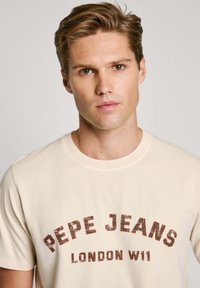 Šviesiai ruda marškinėliai su klasikiniu apvaliu kaklu, turinčiu apvalią, nusidėvėjusią grafiką tamsiai raudona spalva: "PEPE JEANS LONDON W11."