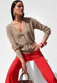 Beige geribbelde cardigan met een V-hals, zwarte knopen, gedragen met felrode broek. Gecombineerd met een rode armband en een bruine handtas.