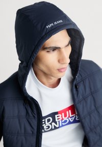 Granatowa pikowana kurtka z kapturem, z logo "PEPE JEANS" z przodu. Nosi na sobie białą koszulę z graficznym nadrukiem.