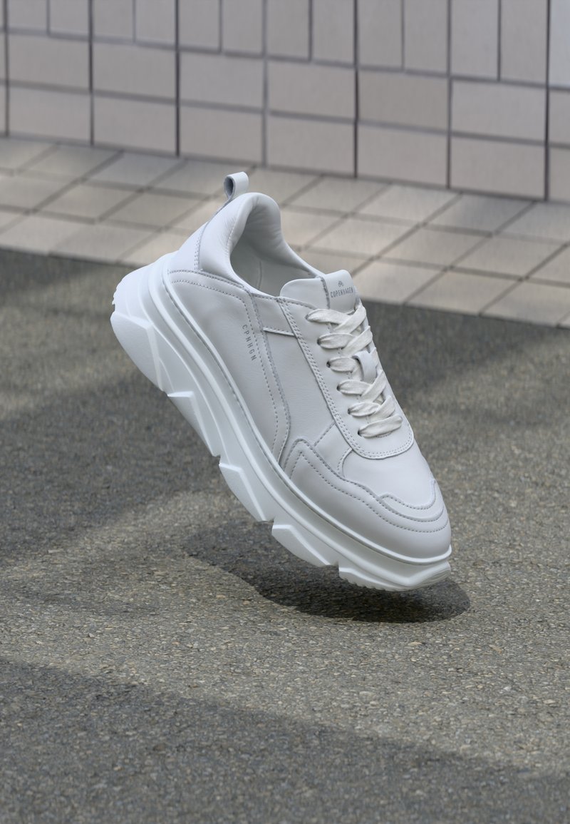 Baskets blanches épaisses à lacets, semelle texturée et détails de marque, placées au-dessus d'une surface pavée près d'un mur carrelé.