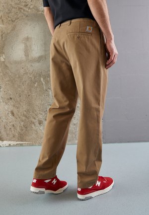 Person iført beige Carhartt bukser og røde New Balance sneakers stående mod en tekstureret beton- og grå væg.