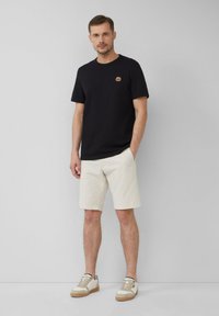 Czarny bawełniany t-shirt z małym haftowanym wzorem, w zestawie z jasnobrązowymi szortami oraz beżowymi trampkami z brązowymi podeszwami i sznurowanym zapięciem.