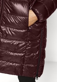 Main dans la poche latérale d'une doudoune matelassée de couleur bordeaux foncé avec une fermeture éclair noire visible le long de la couture latérale.