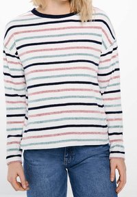 Kobieta w białym swetrze w poziome różowe, zielone i granatowe paski oraz w niebieskich dżinsach, stojąca z rękami w luźnej pozycji.