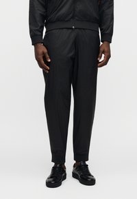 Μαύρο αθλητικό φόρμα από υφασμάτινη υφή με διακριτικό πλέγμα και ελαστικές μανσέτες, σε συνδυασμό με μαύρα sneakers με κορδόνια.