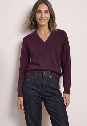 Femme aux cheveux bruns mi-longs portant un pull violet à col en V et un jean bleu foncé, debout contre un mur clair.