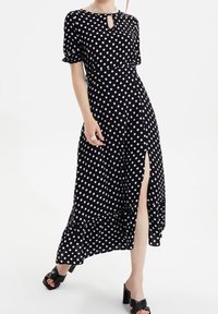 Robe noire à pois avec des manches courtes, décolleté à trou de serrure, fente sur le côté et ourlet à volants. Tissu lisse et partie supérieure ajustée.