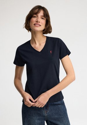 Kobieta o krótkich brązowych włosach, ubrana w granatowy T-shirt z dekoltem w serek i małym czerwonym logo na piersi oraz niebieskie dżinsy, stojąca na jednolitym tle.