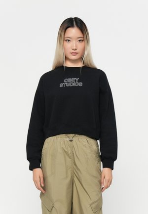 Svart cropped hettegenser med "OBEY STUDIOS" brodert i hvitt på brystet. Har rund halslinning og lange ermer.