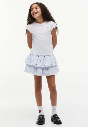 Dziewczyna z długimi kręconymi włosami uśmiechająca się, ubrana w białą koszulkę, spódnicę warstwową w gwiazdy, białe skarpetki w paski i czarne mokasyny, stojąca.