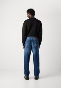 Man met korte dreadlocks, gekleed in een zwart sweatshirt, blauwe spijkerbroek en zwarte schoenen, staand tegenover een effen witte muur.