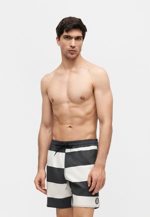 Άντρας χωρίς μπλούζα με κοντά σκούρα μαλλιά, φοράει μαγιό με ασπρόμαυρες ρίγες, στέκεται μπροστά σε απλό λευκό φόντο.