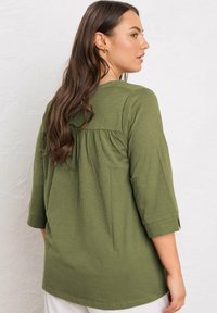 Donna con lunghi capelli castani, che indossa una maglietta verde oliva larga con maniche a tre quarti, girata leggermente di spalle contro un muro bianco testurizzato.