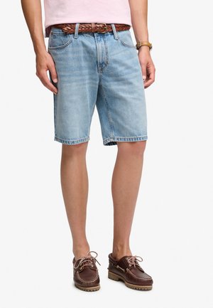Man draagt lichtblauwe denimshorts gecombineerd met een bruine leren gevlochten riem en bruine leren veterschoenen op een witte achtergrond.