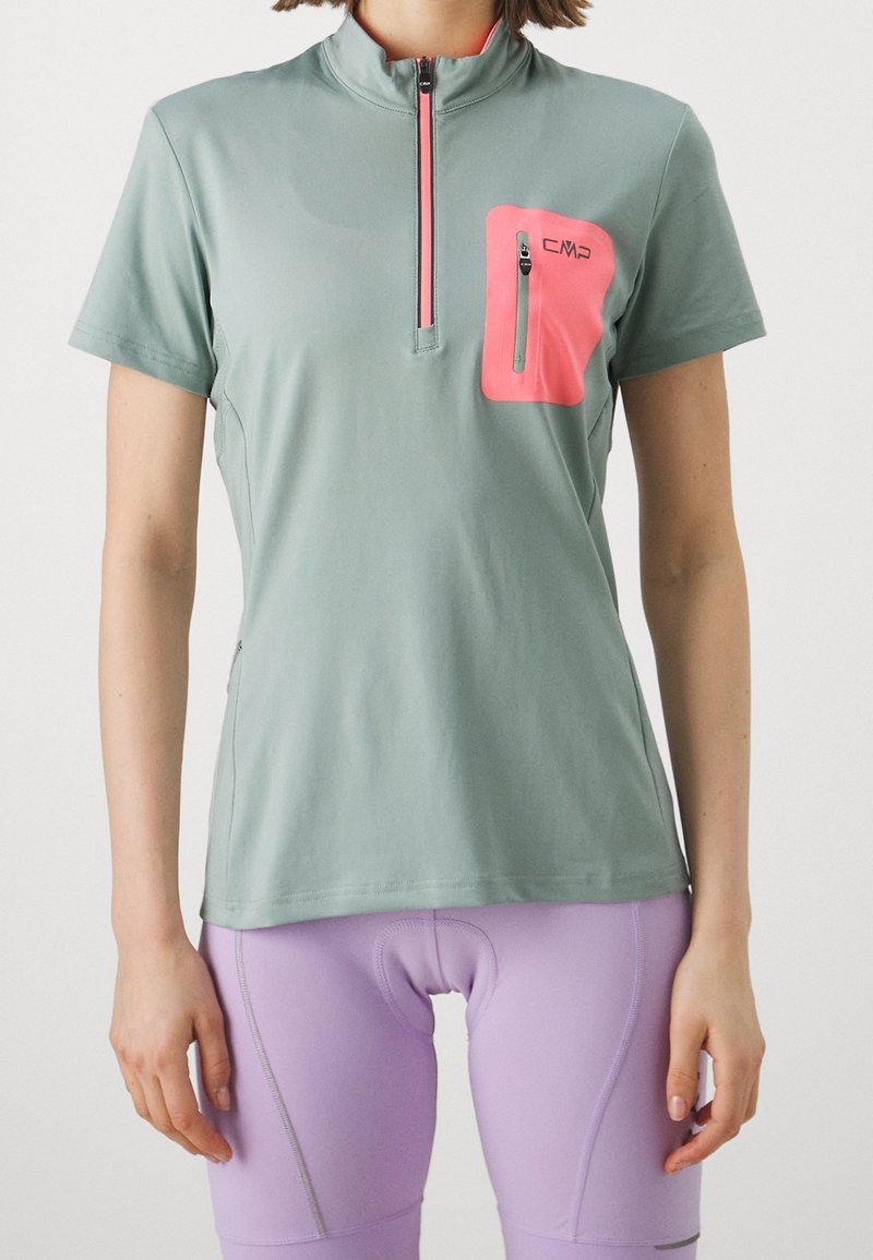 Femme portant un maillot de sport vert à manches courtes avec une poche poitrine zippée rose et un short de cyclisme violet clair.