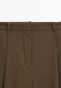 Close-up van de tailleband van een bruine broek met riemlussen en details van de voorknaad op een effen achtergrond.