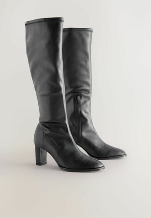 Térdmagasságú fekete bőrcipők hegyes orral, oldali cipzárral és tömbsarkakkal. Simább textúra, varrott részletekkel.