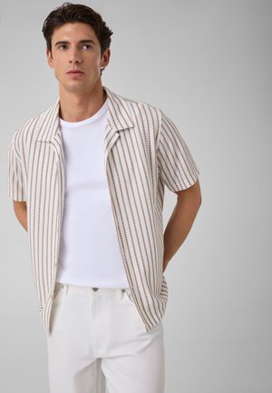Jeune homme portant une chemise ouverte à manches courtes beige et blanche rayée sur un t-shirt blanc avec un pantalon blanc sur un fond gris uni.