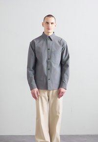 Gray button-up shirt with a chest pocket, paired with loose beige trousers. Smooth fabric with a structured fit and pointed collar.