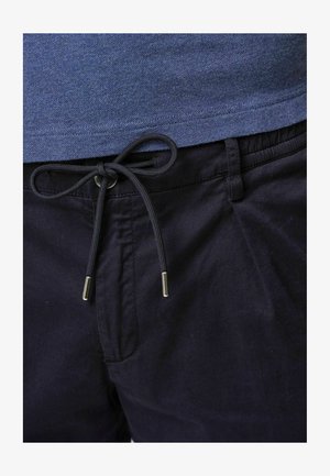 Pantalones cortos de algodón azul marino con cintura ajustable y puntas de metal plateadas, exhibidos sobre una camisa azul. Tejido texturizado visible.