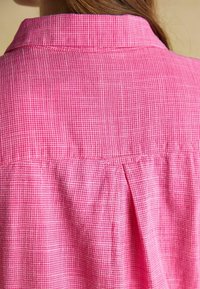 Plano detalle de la parte superior de la espalda de una persona que lleva una camisa de cuello con pequeños cuadros rosas y blancos con un pliegue central en la espalda.