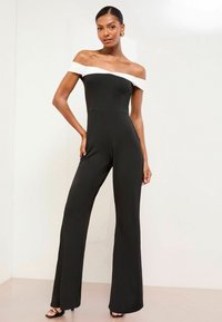 Jumpsuit negro sin mangas con un cuello blanco, cuerpo ajustado y pantalones de pierna ancha, hecho de una tela suave. La modelo posa contra un fondo neutro.
