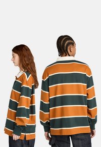 Dwóch ludzi w pomarańczowych i ciemnozielonych poziomych pasach na koszulach rugby, pokazanych z boku i z tyłu na jednolitym tle.