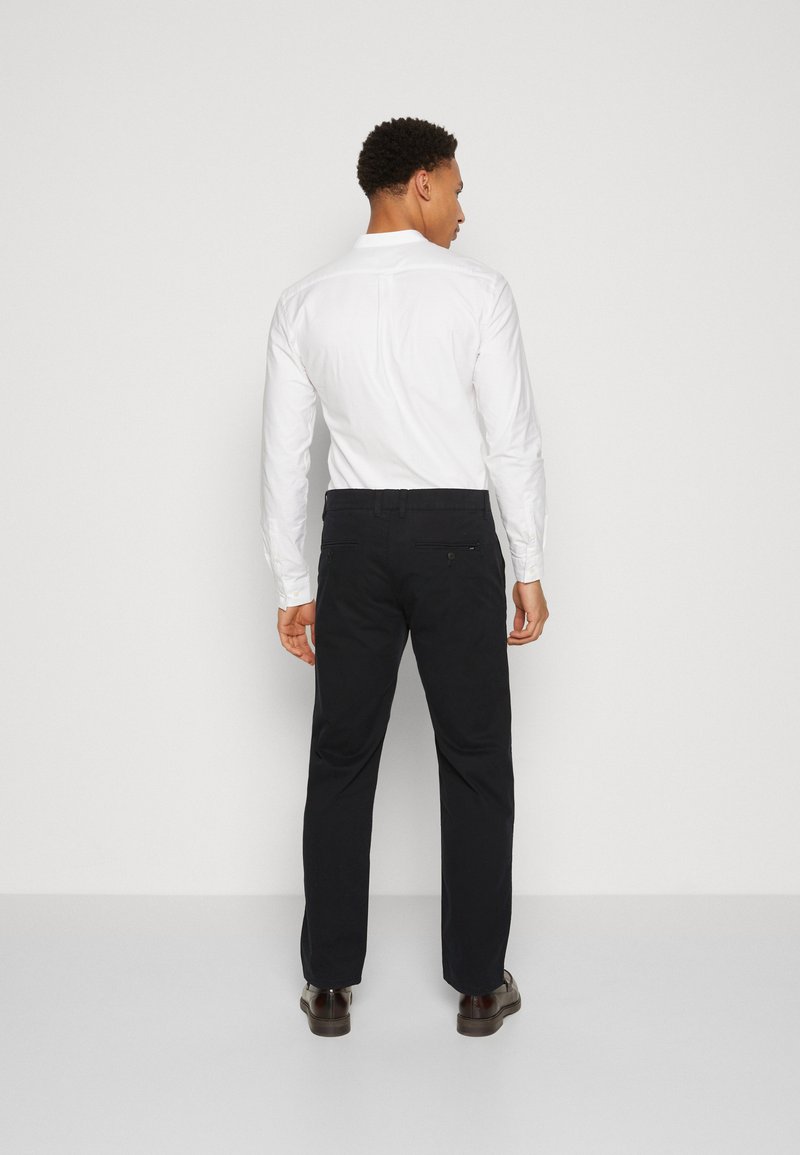 Tenue masculine comprenant une chemise blanche à boutons et un pantalon chino noir, mettant en valeur une texture lisse et un design ajusté, vue de dos avec des chaussures brunes.