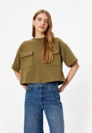 Olivgrünes, kurzes T-Shirt aus geripptem Material mit zwei Brusttaschen; kombiniert mit hochgeschnittenen, geraden blauen Jeans.