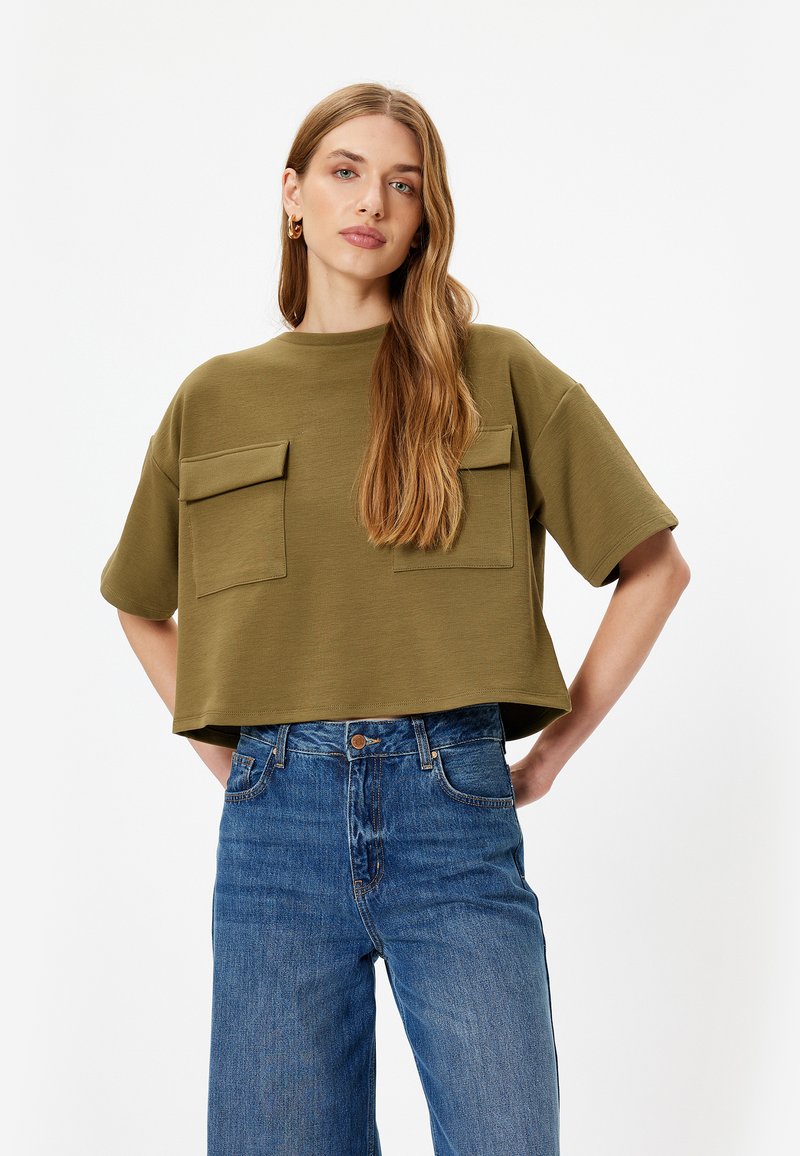 Olijfgroen, cropped t-shirt in ribbeltextuur met twee borstzakken; gedragen met hoog-gerimpelde, rechte spijkerbroek in blauw.