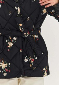 Prešívaná čierna bunda s kvetinovou potlačou, zaviazaná v páse zladeným opaskom, nosená na svetlých nohaviciach.