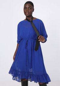 Vloeibare blauwe chiffon jurk met een ronde halslijn, elastische taille en gerimpelde zoom. Gestyled met een zwarte strikaccessoire en kniehoge zwarte laarzen.