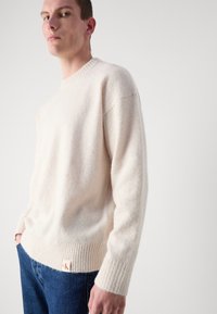 Kremfarget strikket genser med ribbestrikket nederkant og mansjetter. Har rund hals og en fremtredende logoetikett nederst til venstre. Båret med blå jeans.