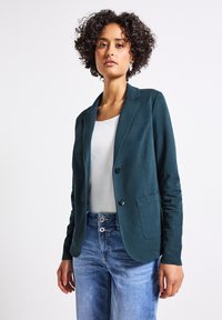 Teal sako vyrobené z měkké látky s klopovým límcem, zapínáním na jeden knoflík a dvěma předními kapsami, nošené přes bílý top a džíny.