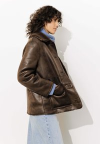 Veste en cuir marron avec un devant à boutons, de grandes poches et un col en fourrure. Portée sur un pull bleu clair et un jean en denim.