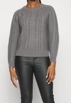 Kvinde iført en grå kabelstrikket sweater og sorte læderbukser står mod en ensfarvet lys baggrund.