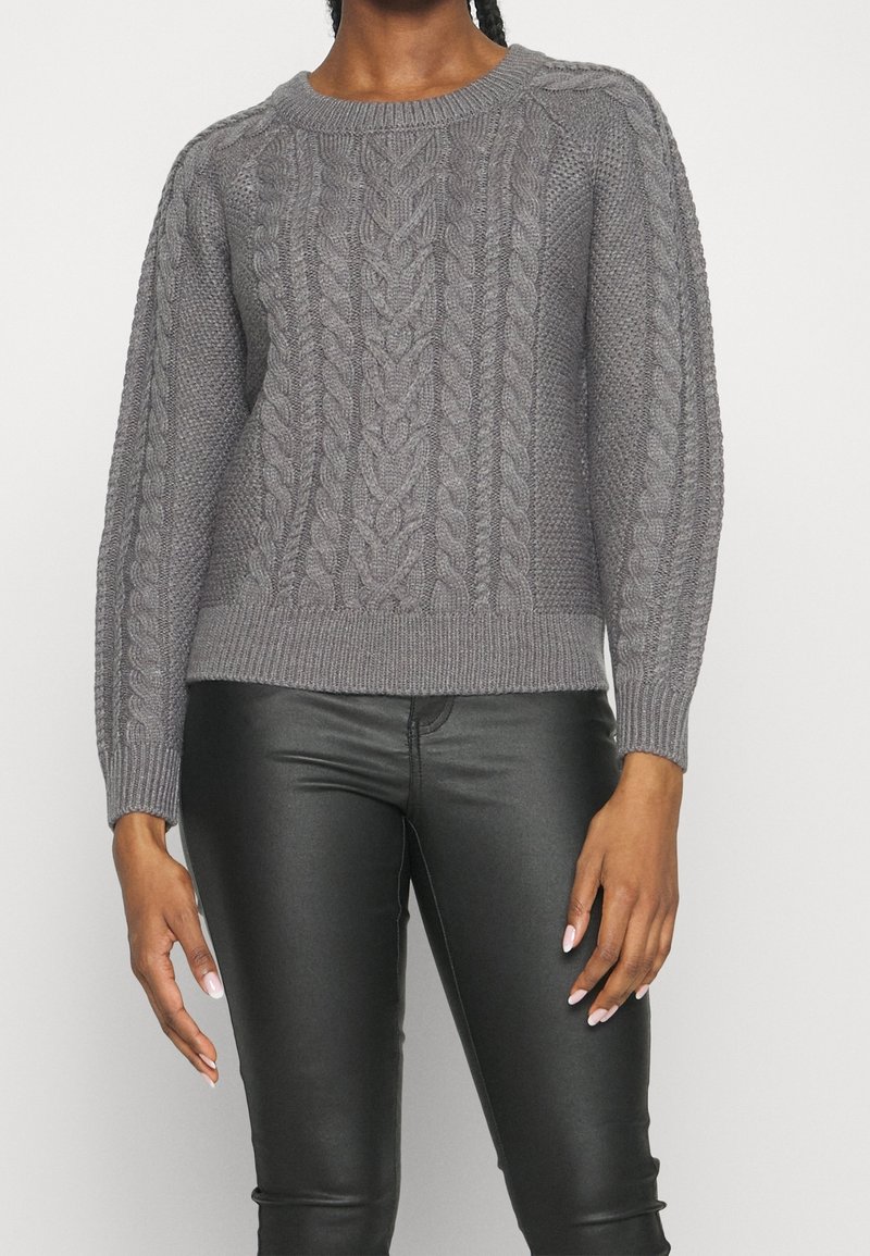Femme portant un pull gris à torsades et un pantalon en cuir noir, debout devant un fond clair uni.